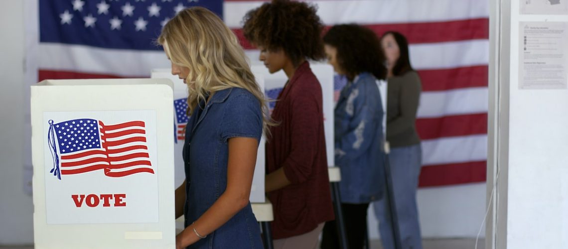 black vote, Four women of various demographics, young blonde woman in front, filling in ballots and casting votes in booths at polling station, US flag on wall at back. Focus on booth signage