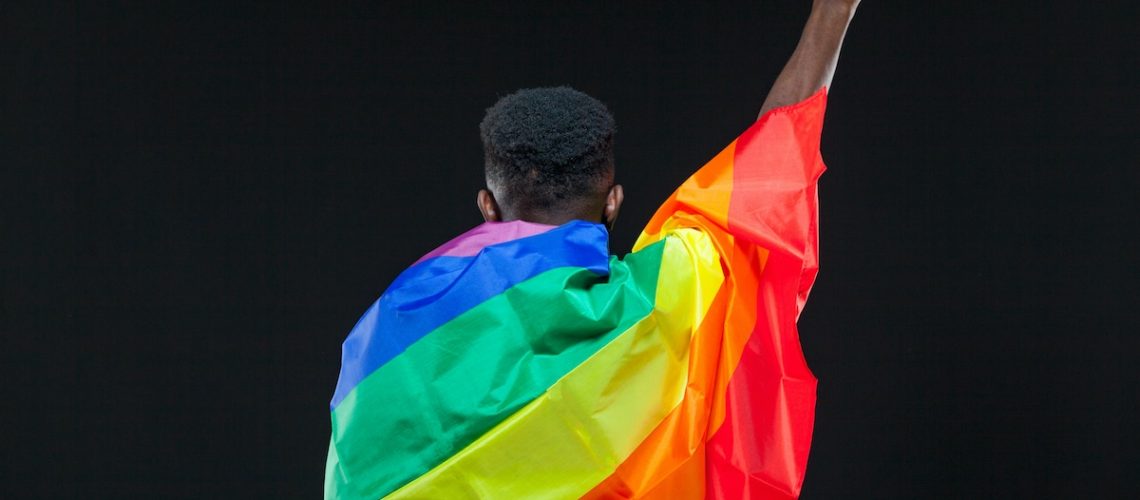 Back view of young african american man wrapped in a rainbow flag standing with raised fist isolated on black background. Concept of The LGBT community, minority rights, protection of human rights