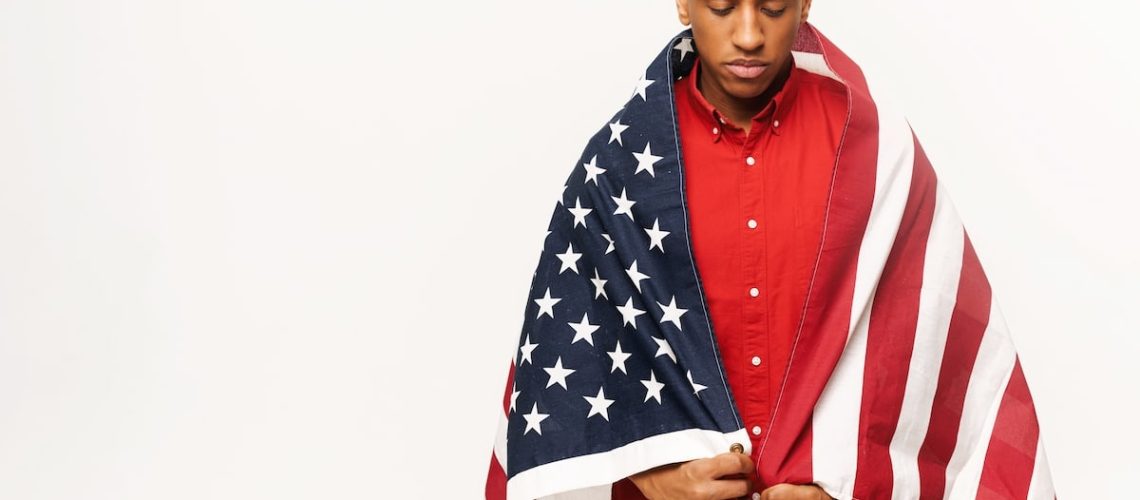 Sad multiracial man defends his freedom and equal rights. Serious worried African-American guy stands covered with american flag isolated over white background