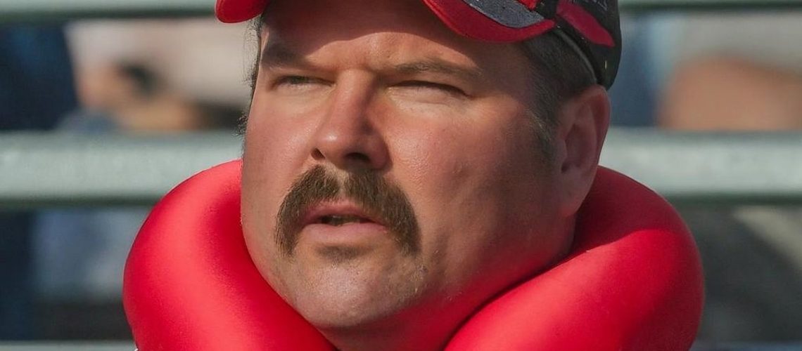 A realistic photograph OF A 40 YEAR OLD MALE AMERICAN stereotypical country NASCAR FAN WITH A BRUSH MOUSTACHE SITTING IN THE STANDS OF A FORMULA E RACE TRACK, WEARING A RED TRAVEL NECK PILLOW